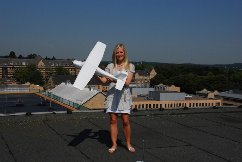 Ein blondes Mädchen steht auf einem Flachdach und hält ein weißes kleines Flugzeug in den Händen.