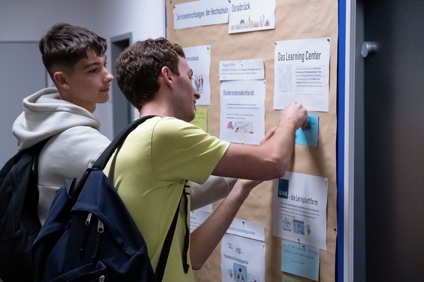 Zwei Männer pinnen einen Zettel auf eine Pinnwand, an der ein großes, braunes Plakat hängt. Auf dem Plakat pinnen schon andere weiße und blaue Zettel, auf denen etwas geschrieben steht.. 