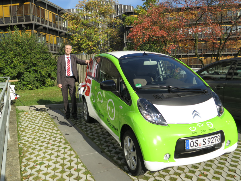 Elektroauto Auf dem Bild sieht man einen Mann neben einem bunt beklebten Kleinwagen stehen. Zum Auto führt ein dickes Stromkabel.