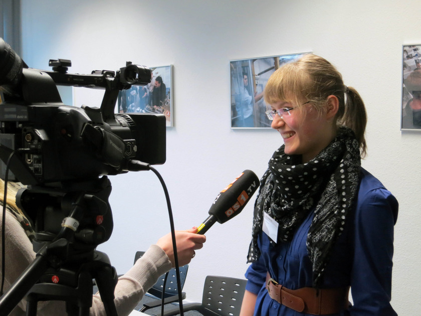 Auf dem Bild sieht man, wie ein junges blondes Mädchen mit Brille vor laufender Kamera ein Interview gibt.