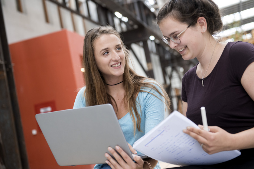 Zwei Studentinnen sitzen nebeneinander und haben einen Laptop und Papiere in der Hand.