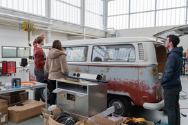 Drei Personen stehen in einer Werkstatt neben einem alten, rostigen VW-Bus mit Solarpanelen auf dem Dach und besprechen ein Dachsolarkonzept.