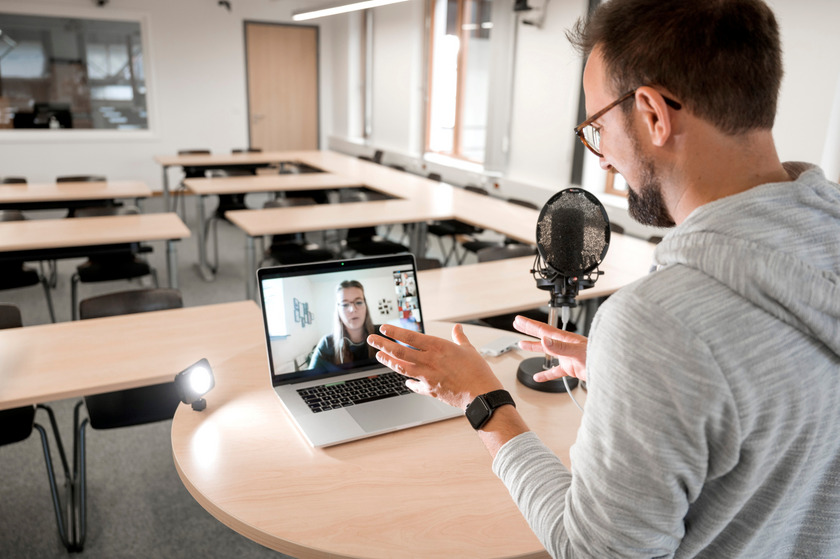 Das Bild zeigt einen jungen Lehrenden in einem leeren Seminarraum vor einem Laptop in einer Videokonferenz.