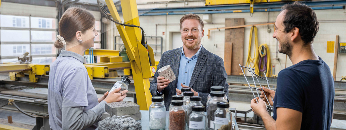 Professor bespricht in einer Werkhalle mit zwei Personen Materialproben.