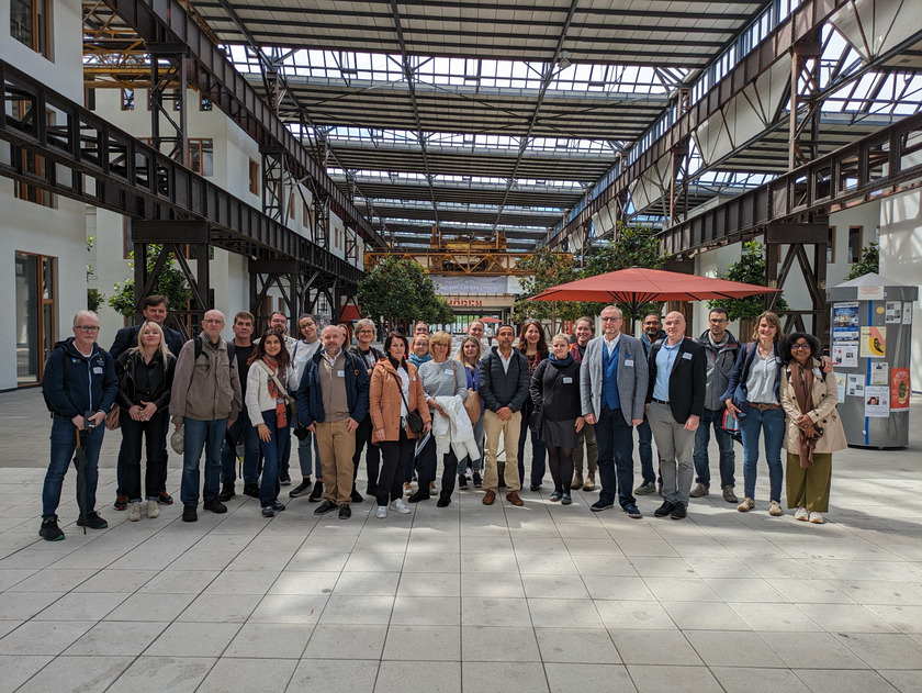 Eine große Gruppe von Menschen, die in einer geräumigen, industriell gestalteten Halle stehen. Die Halle hat hohe Decken mit Stahlträgern und großen Fenstern, die viel Licht hereinlassen. Die Gruppe posiert für ein Gruppenfoto, viele der Teilnehmer tragen Namensschilder. Im Hintergrund sind Pflanzen und Informationsstände zu sehen. Die Atmosphäre wirkt professionell und kollegial, und die Halle scheint ein Veranstaltungsort für größere Treffen oder Konferenzen zu sein.