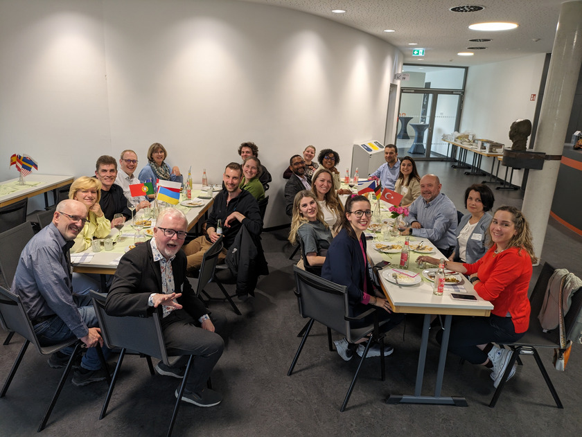 Eine Gruppe von Menschen, die um zwei lange Tische sitzen und eine Mahlzeit genießen. Die Tische sind festlich gedeckt, und die Teilnehmer lachen und unterhalten sich angeregt. Einige der Gäste halten kleine Landesflaggen, die auf dem Tisch stehen. Die Atmosphäre ist fröhlich und gesellig. Der Raum hat eine moderne Einrichtung, und im Hintergrund ist ein offener Bereich zu sehen, der möglicherweise als Buffetbereich dient.