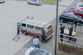 Sechs Personen montieren auf einem alten weißen VW-Bus mit Roststellen ein Metallgestell auf dem Dach auf einem Parkplatz