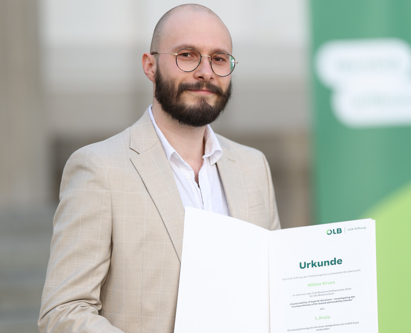 Ein junger Mann hält eine Urkunde in der Hand und lächelt in die Kamera.