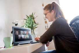 Das Bild zeigt eine junge Frau, die zuhause an ihrem Schreibtisch sitzt und auf ihren Laptop schaut, auf dem mehrere Personen in einer Videokonferenz miteinander sprechen.