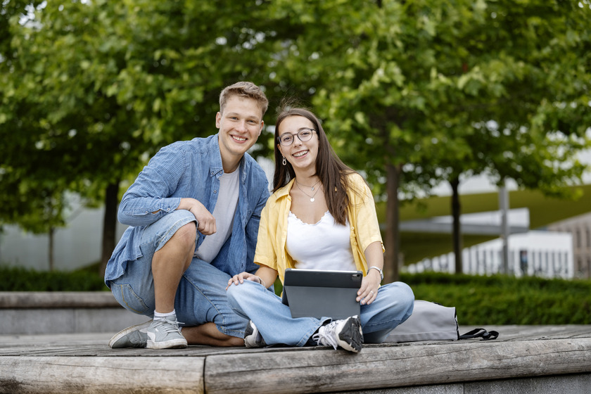 Zwei Studierende sitzen auf einer Bank und lachen in die Kamera.