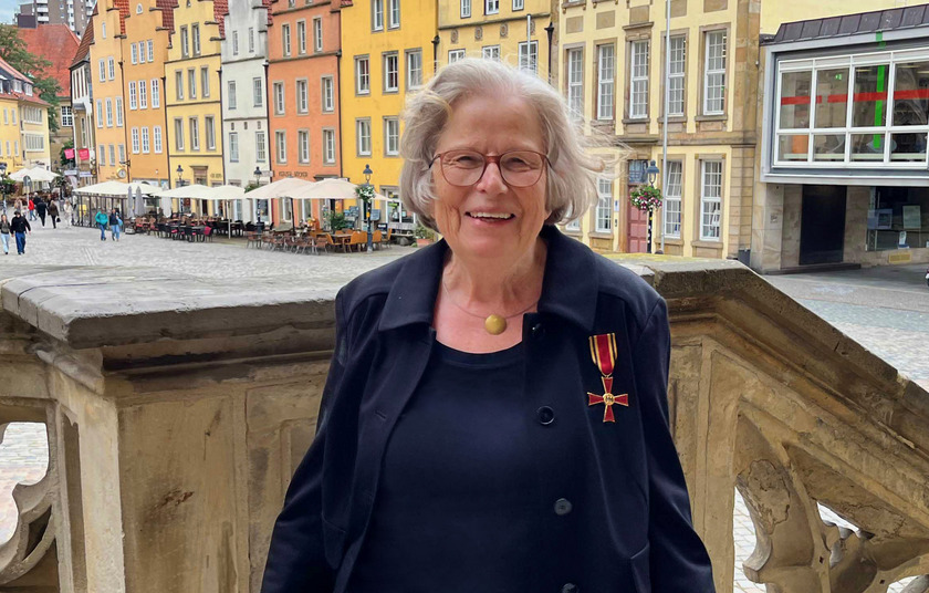 Eine ältere Dame steht auf der Rathaustreppe vor den alten Osnabrücker Giebellhäusern. Sie trägt an ihrem Blazer das rot-goldene Bundesverdienstkreuz und lächelt in die Kamera.