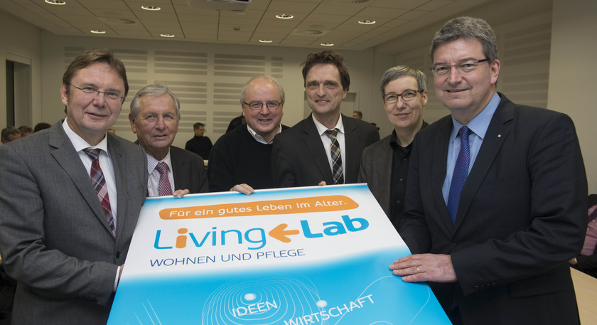 Man sieht sechs Personen in Anzügen, die ein Plakat mit den Wörtern "Living Lab" in der Hand halten.