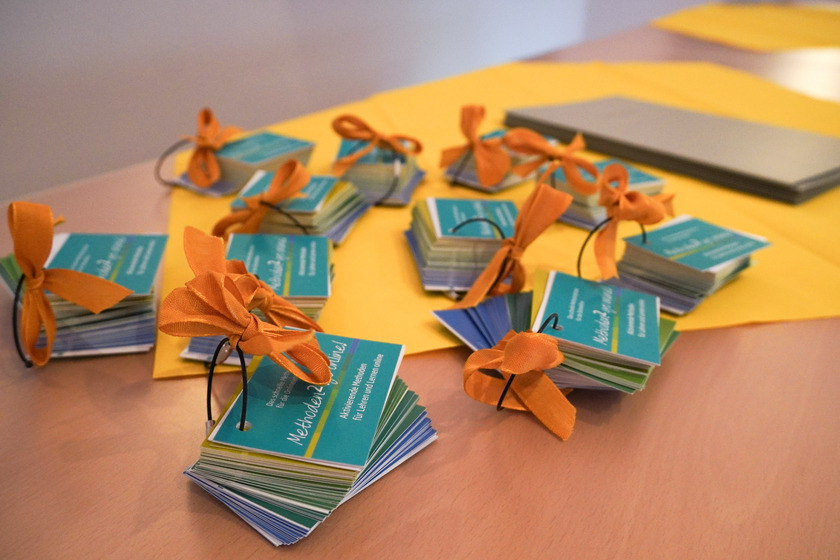 Auf einem Tisch liegen zehn Kartenstapel, die jeweil zusammengebunden sind zu einem Dossier. Sie sind blau gefärbt und an dem Band, dass die Dossiers zusammenhält, ist eine orangefarbene Schleife gebunden. 