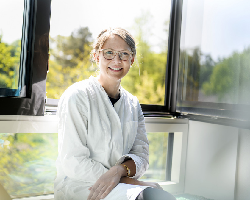 Eine junge Frau im weißen Laborkittel sitzt im Labor am offenen Fenster und lächelt in die Kamera.