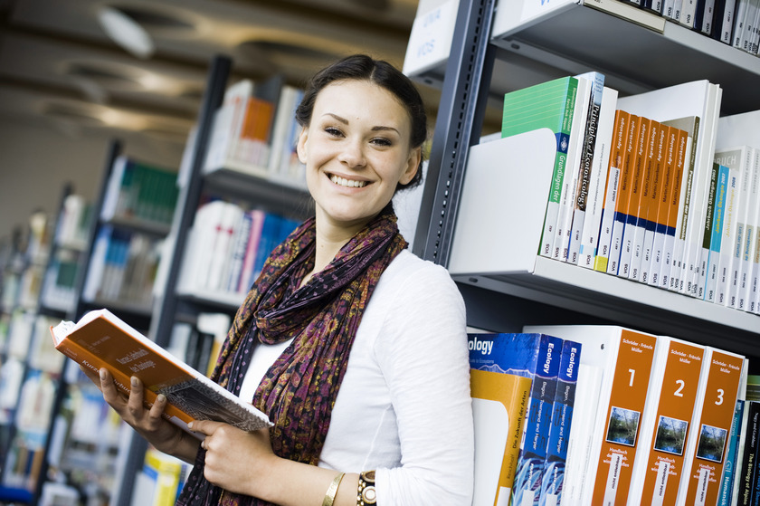 Auf dem Bild sieht man eine junge Frau, die vor einem Bücherregal steht.