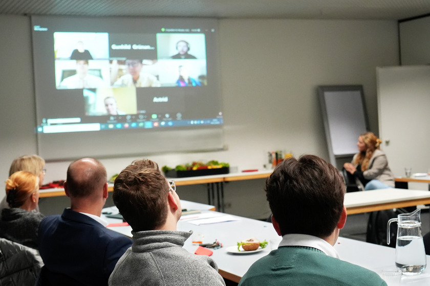 In einem Konferenzraum verfolgen mehrere Personen eine Online-Präsentation auf einer großen Leinwand. Die Leinwand zeigt einen Zoom-Call mit verschiedenen Teilnehmer*innen, deren Videobilder in einer Galerie-Ansicht angezeigt werden. Es sind mehrere Gesichter sichtbar, jedoch sind die Details der Personen unscharf. Einige der Teilnehmenden haben ihre Videokameras aktiviert, während andere nur ihre Namen eingeblendet haben.  Im Vordergrund sieht man den Rücken von mehreren Personen, die auf Stühlen sitzen. Eine Person trägt eine grüne Jacke, während eine andere Person eine graue Fleecejacke trägt. Die Köpfe der Zuhörer sind in Richtung der Leinwand gedreht. Auf dem Tisch befinden sich Notizen, ein Teller mit einem belegten Brötchen, ein Glas Wasser und ein Krug mit Wasser.  Im Hintergrund sitzt eine Frau mit blonden Haaren und schwarzer Hose auf einem Stuhl, neben einer Tafel oder einem Flipchart. An den Wänden des Raumes sind Dekorationen und verschiedene Büromaterialien zu sehen. Der Raum ist gut beleuchtet, und es herrscht eine aufmerksame Atmosphäre.