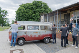 Mehrere Personen fotografieren und betrachten einen alten, rot-weißen VW-Bus mit Dachträger vor einem Gebäude mit überdachter Terrasse