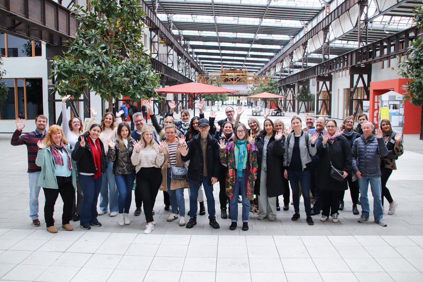 Eine Gruppe von rund 25 Personen steht in einer großen, hellen Halle und winkt lächelnd in die Kamera. Im Hintergrund sind Bäume in Pflanzkübeln, Sitzgruppen mit roten Sonnenschirmen und eine industrielle Stahlkonstruktion zu sehen.