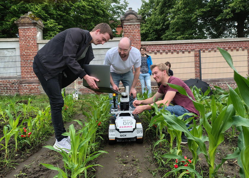 Drei Studierende stehen mit einem kleinen Roboter auf einem Feld.
