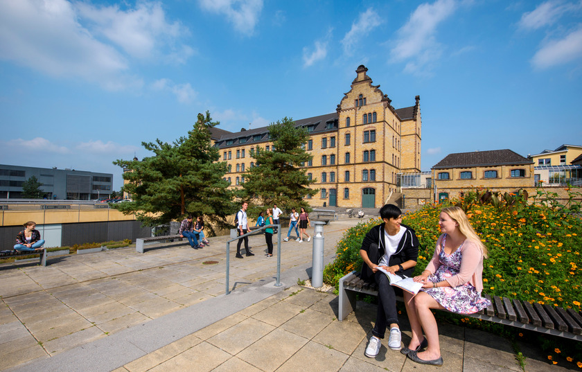 Studierende befinden sich auf dem Caprivi Campus. Im Hintergrund ist ein altes Kasernengebäude zu sehen und im Vordergrund stehen und sitzen Studierende. 