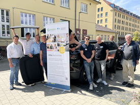 9 Männer stehen zum Abschluss der Veranstaltung vor einem PKW und einem Rollup.