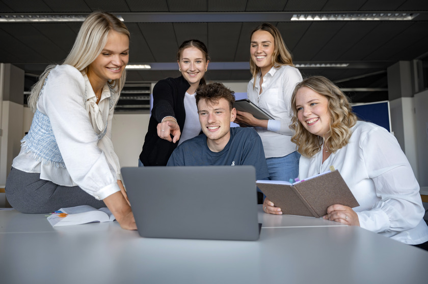 Fünf Studierende sitzen und stehen um einen Laptop herum und erledigen eine Gruppenarbeit.