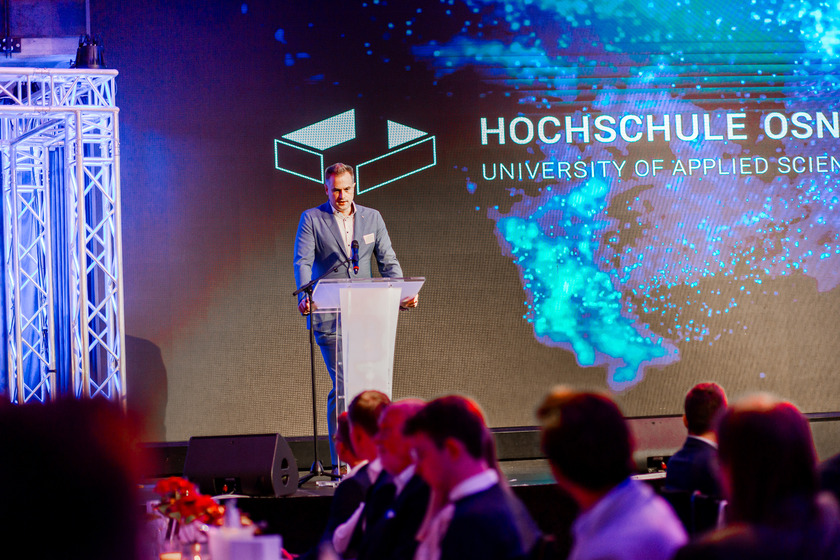 Ein Mann in grauem Anzug steht hinter einem Rednerpult. Im Hintergrund ist das Logo der Hochschule Osnabrück zu sehen. 