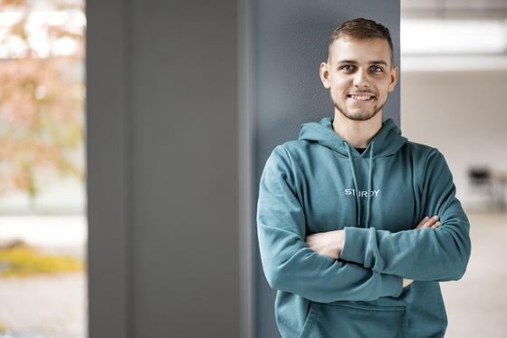 Ein junger Mann, der lächelnd in die Kamera blickt. Er steht lehnend an einer grauen Säule in einem offenen Raum mit einem unscharfen Hintergrund, der Herbstbäume und ein helles Gebäude zeigt. Der Mann hat kurze, hellbraune Haare und trägt einen türkisfarbenen Hoodie mit der Aufschrift "STURDY" auf der Brust. Seine Arme sind gekreuzt, was eine selbstsichere Haltung suggeriert. Sein freundliches Auftreten und die entspannte Körperhaltung vermitteln ein Gefühl von Zuversicht und Offenheit.