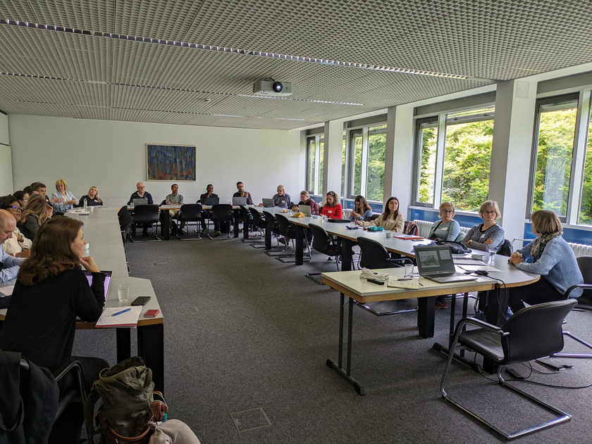 Eine Gruppe von Menschen, die in einem Konferenzraum an einem langen Tisch sitzen. Der Raum ist hell erleuchtet, mit großen Fenstern auf der rechten Seite, die viel Tageslicht hereinlassen. Die Teilnehmer sitzen an drei Seiten des Tisches und blicken auf eine Person, die vorne spricht. Die Stimmung wirkt konzentriert und professionell, einige Teilnehmer haben Laptops vor sich, während andere Notizen machen. Ein Gemälde hängt an der Wand im Hintergrund, und der Raum hat einen Teppichboden.