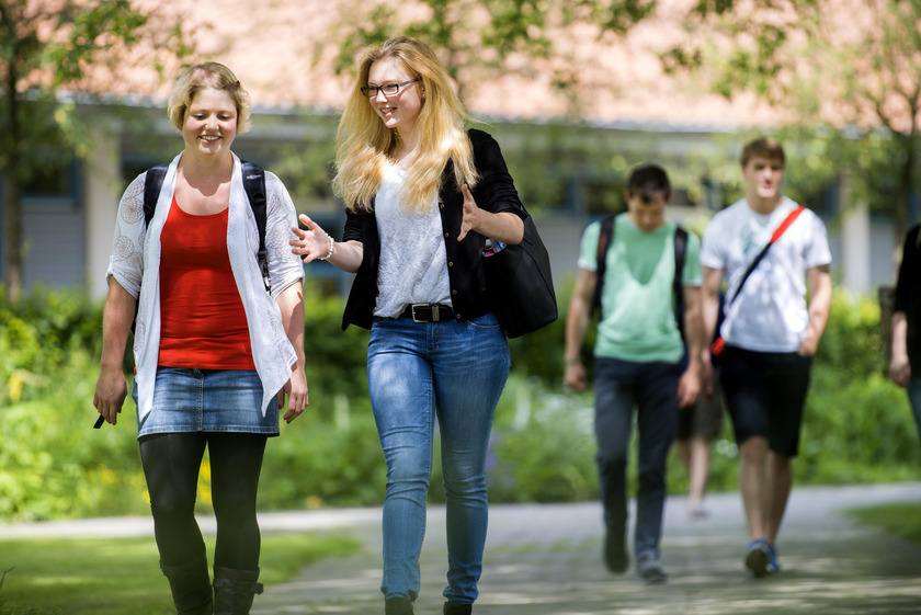 Das Bild zeigt Studierende, die über einen grünen Campus gehen und im Gespräch vertieft sind.