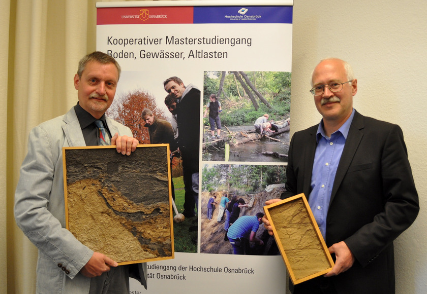 Das Bild zeigt zwei Männer, die eingerahmte Bodenschichten in der Hand halten.