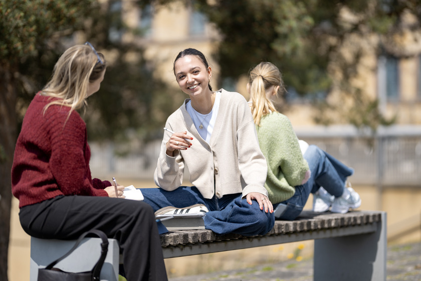 Drei Studentinnen sitzen draußen auf einer Bank. Eine von ihnen hält einen Stift in der Hand und lächelt direkt in die Kamera.