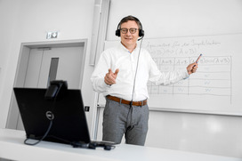 Das Bild zeigt einen Mann mittleren Alters, der mit einem Headset vor einem Laptop steht und auf das Whiteboard hinter sich zeigt.