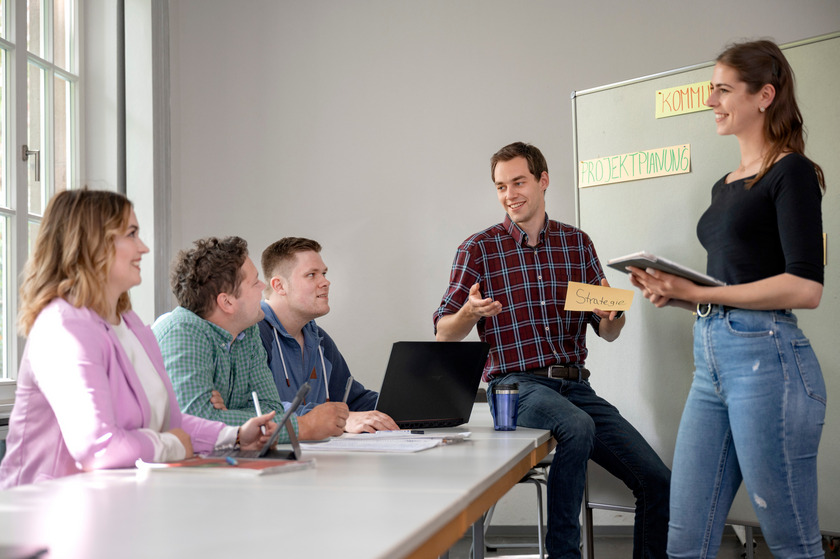 Fünf Studierende unterhalten sich im Rahmen einer Gruppenarbeit und halten ihre Ergebnisse an einer Pinnwand fest.