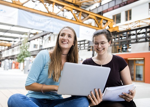 Zwei Studentinnen sitzen in der Halle des Campus Lingen und haben einen Laptop oder einen Block in der Hand.