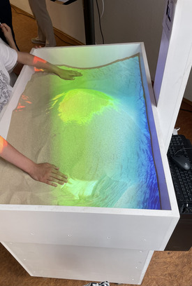 Bild einer mit Sand gefüllten Holzbox, der farbig beleuchtet ist.