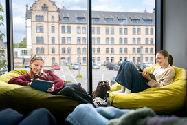 Zwei Studierende sitzen in der Lernlandschaft der Caprivi Lounge am Caprivi Campus in Sitzsäcken und arbeiten mit ihren Tablets. Im Hintergrund ist ein altes Kasernengebäude zu sehen.