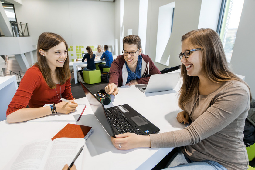 Das Bild zeigt junge Menschen, die an einem Tisch sitzen und mit einem Laptop arbeiten.