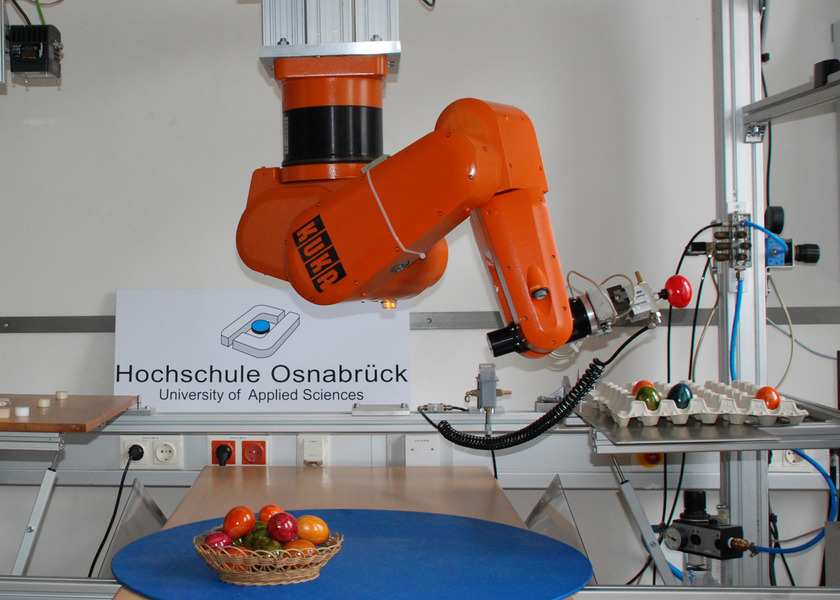 Das Bild zeigt einen orangenen Robotergreifarm, der bunte Ostereier aus einem Nest in einen Eierkarton legt.