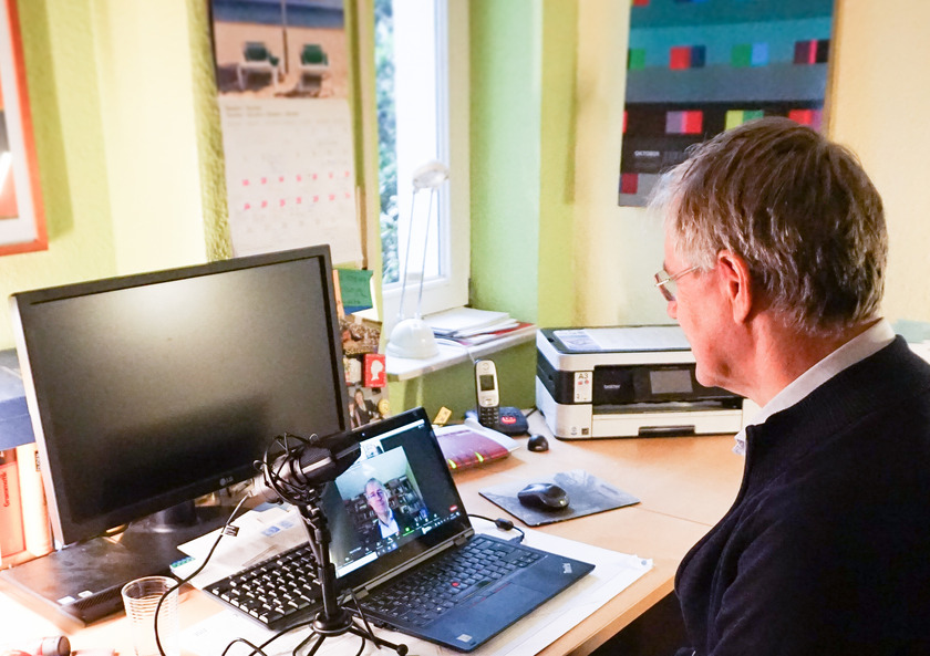 Prof. Dr. Peter Mayer sitzt an seinem Schreibtisch, vor ihm steht sein Laptop und ein Mikrofon. Er unterhälts sich über einen Videochat mit den Teilnehmenden.